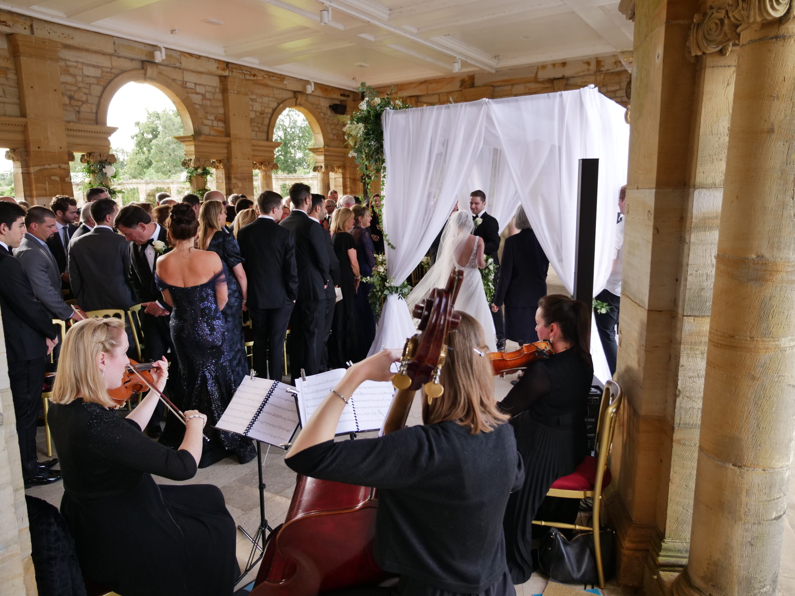 Exclusive String Trio of 2 violins and double bass playing for a wedding ceremony at Hever Castle, The Loggia. The bride and groom are facing each other under a white net awning and the guests are standing.